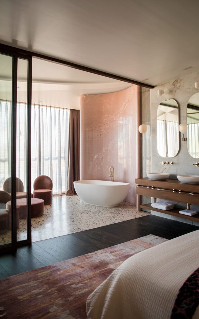 A photo of a chic mid-century modern bedroom with an open-plan ensuite. The bedroom has a dark-stained wood floor and a plush area rug. There's a freestanding oval tub near the sleeping area, framed by a curved partial wall clad in blush pink marble. A walnut floating vanity with double sinks and oval mirrors is placed near the tub. The bathroom has a terrazzo floor. The shared space includes sculptural lounge seating, abstract wall art, and a touch of velvet in accessories. Large sliding glass doors bring in soft natural light, while sheer curtains add privacy without breaking flow.