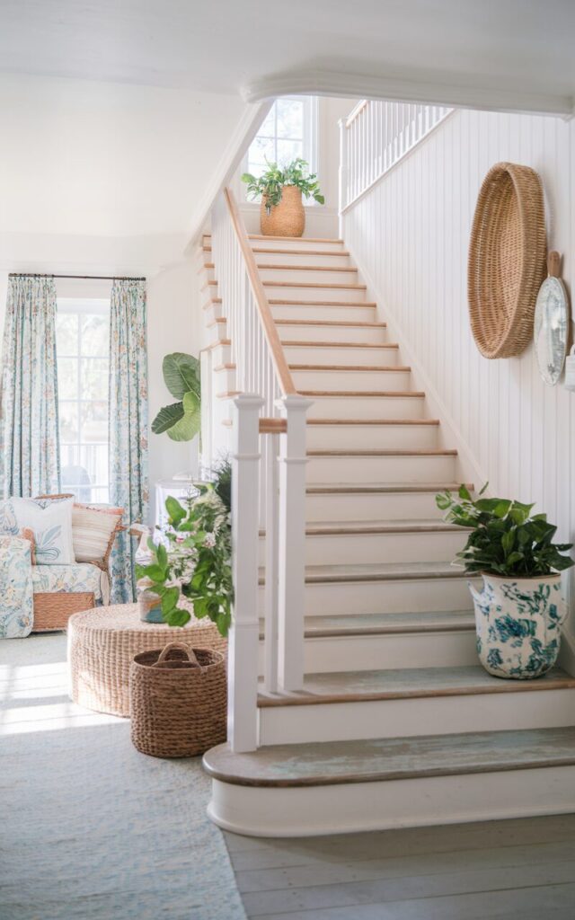 A photo of a bright, airy staircase in a California coastal cottagecore style home. The staircase has open risers that allow light to flow freely between the steps. The treads are weathered wood with a soft white-washed finish, and the railing is simple white. Surrounding the staircase, sunlit rooms have breezy linen curtains, woven baskets, potted greenery, and vintage floral textiles. The overall vibe is fresh, relaxed, and charmingly rustic, blending coastal lightness with cozy cottage warmth.