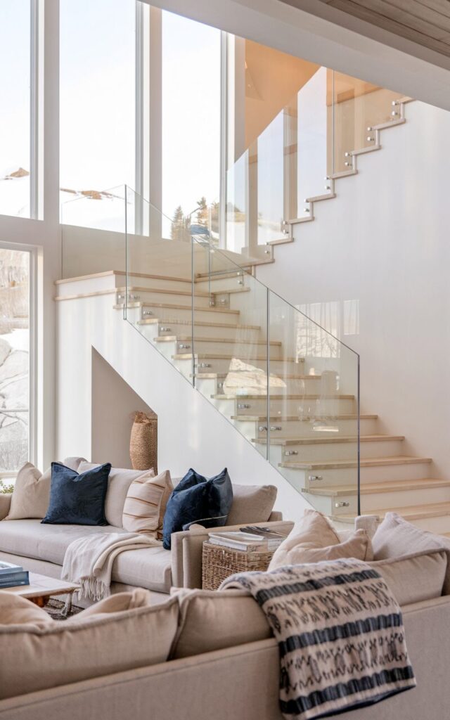 A photo of a living room with an alpine chic and coastal style. There's a floating staircase with sleek glass railings. The stairs have light-toned wooden treads and are supported by a hidden stringer. The glass panels are frameless and reflect soft natural light from large windows. The living room is fully furnished with plush sofas, layered throws, woven textures, and a neutral palette accented with ocean blues.