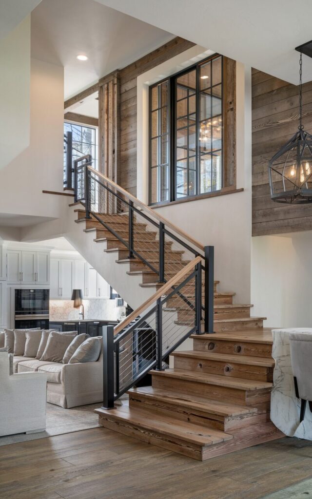 A photo of a modern luxe farmhouse-style house with a staircase positioned between the living room and kitchen. The stairs have rich reclaimed wood treads with natural knots and weathered textures, exuding rustic charm. The railing combines matte black metal with wooden handrails, blending farmhouse warmth with modern sophistication. The surrounding spaces are elegantly furnished with plush sofas, industrial-style lighting, and sleek kitchen cabinetry with marble countertops. Soft natural light filters through large windows, highlighting the beautiful contrast between rugged wood and luxe finishes.