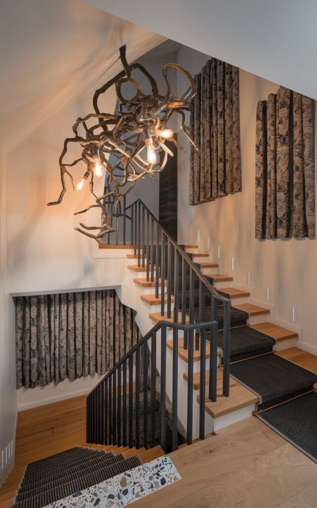 A photo of a modern eclectic home interior with a dramatic sculptural chandelier mounted above the staircase landing. The chandelier has an artistic arrangement of intertwined metal branches with exposed Edison bulbs. The staircase has natural wood and terrazzo treads with a runner and sleek black railing.