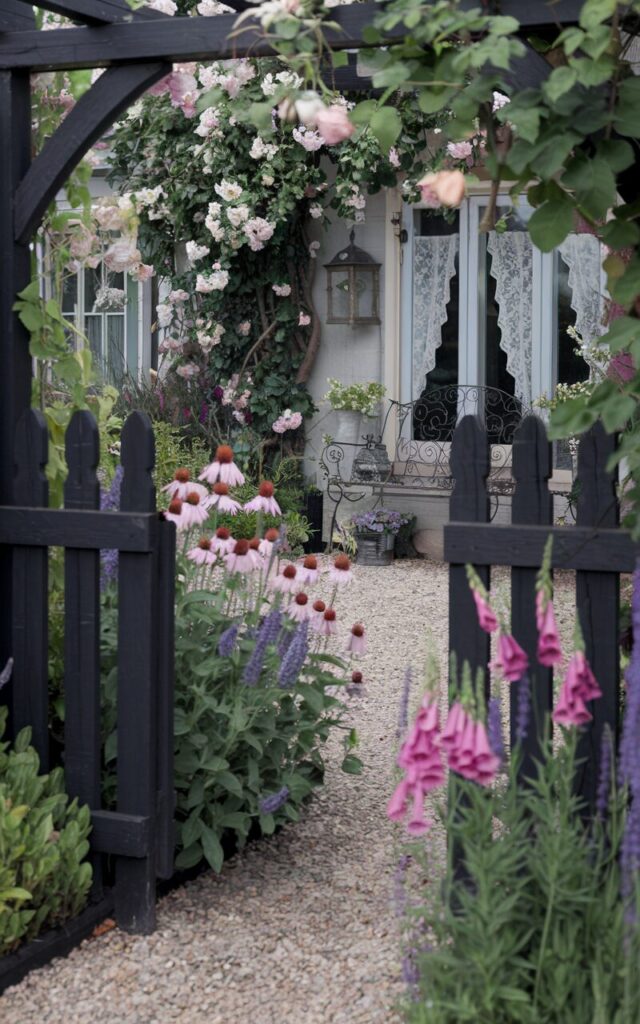 A photo of a cottagecore backyard viewed from outside the picket garden fence. The wooden panels of the fence are painted a deep, moody black. Blooming perennials like echinacea, lavender, and foxglove are planted in a gentle arc, spilling color and texture onto a gravel path. Climbing roses and trailing vines peek over the fence. Beyond the garden, the backyard reveals glimpses of a cozy cottage with lace curtains, hanging lanterns, and a wrought iron bench nestled under a flowering tree.